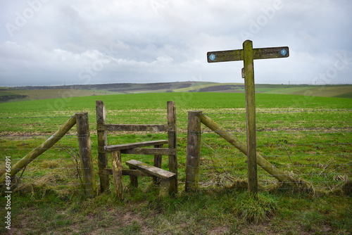 South Downs Way fingerpost next to a footpath stile crossing a fence into a grass meadow under an overcast cloudy sky. The route is a long distance bridleway in Sussex, England, UK.