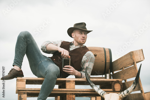 Cowboy wearing hat. Attractive man with whiskey or brandy. Western life. Handsome bearded west farmer. Portrait of man cowboy or farmer. Western.