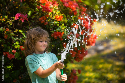 Happy kid boy pours water from a hose. American kids childhood. Child watering flowers in garden. Home gardening.