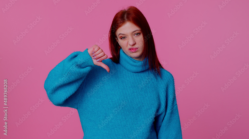 Upset pretty stylish teen girl in blue sweater showing thumbs down sign gesture, expressing discontent, disapproval, dissatisfied, dislike. Young adult woman. Indoor studio shot on pink background Upset pretty stylish teen girl in blue sweater showing thumbs down sign gesture, expressing discontent, disapproval, dissatisfied, dislike. Young adult woman. Indoor studio shot on pink background