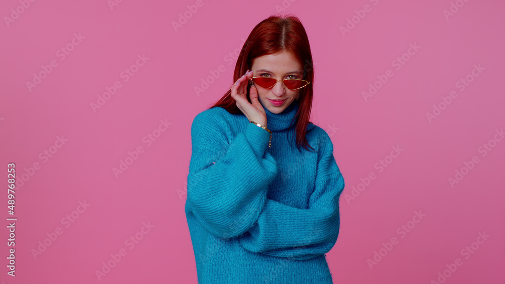 Portrait of seductive cheerful teen redhead girl in blue sweater wearing sunglasses, charming smile. People emotions concept. Young lovely adult woman posing isolated on pink studio wall background Portrait of seductive cheerful teen redhead girl in blue sweater wearing sunglasses, charming smile. People emotions concept. Young lovely adult woman posing isolated on pink studio wall background