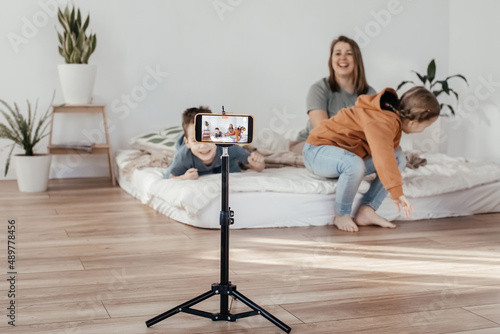 the family smiles and records a video on the phone. bloggers shoots a video clip at home. the phone is on a tripod. family mom and two children are sitting on the bed and posing for the camera