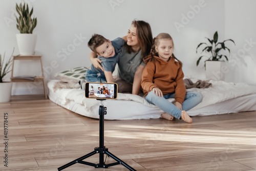 the family smiles and records a video on the phone. bloggers shoots a video clip at home. the phone is on a tripod. family mom and two children are sitting on the bed and posing for the camera