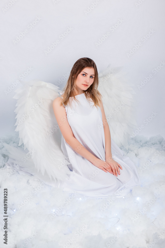 An attractive girl in a white tunic with large white wings behind her back poses while sitting in white luminous clouds. An attractive girl in a white tunic with large white wings behind her back poses while sitting in white luminous clouds.