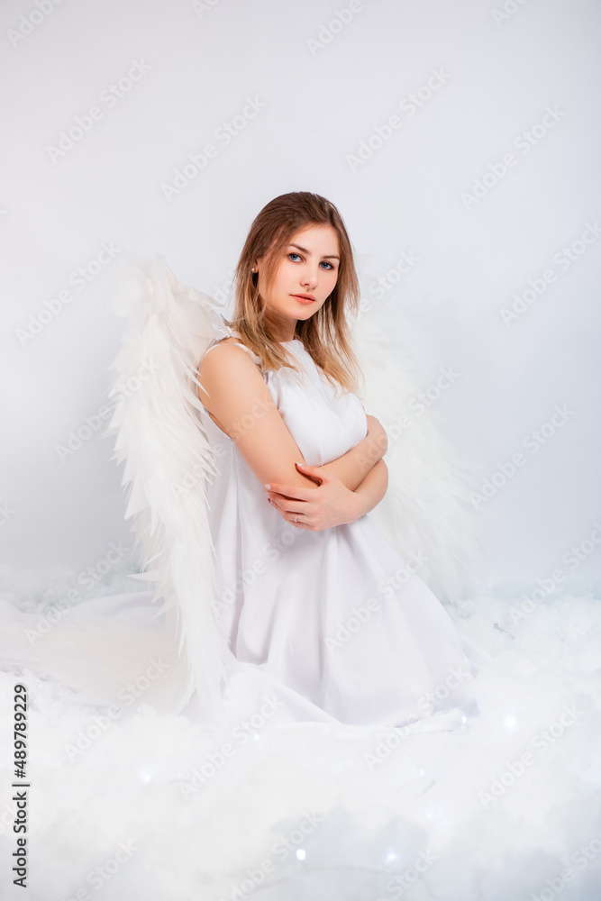 An attractive girl in a white tunic with large white wings behind her back poses while sitting in white luminous clouds. An attractive girl in a white tunic with large white wings behind her back poses while sitting in white luminous clouds.