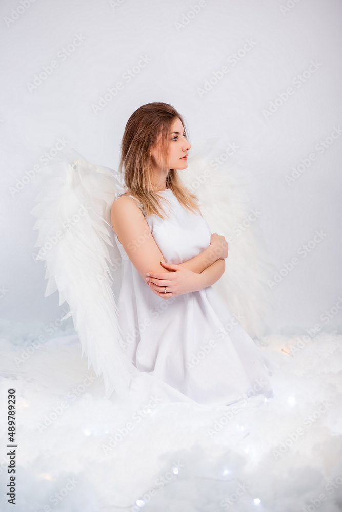 An attractive girl in a white tunic with large white wings behind her back poses while sitting in white luminous clouds. An attractive girl in a white tunic with large white wings behind her back poses while sitting in white luminous clouds.