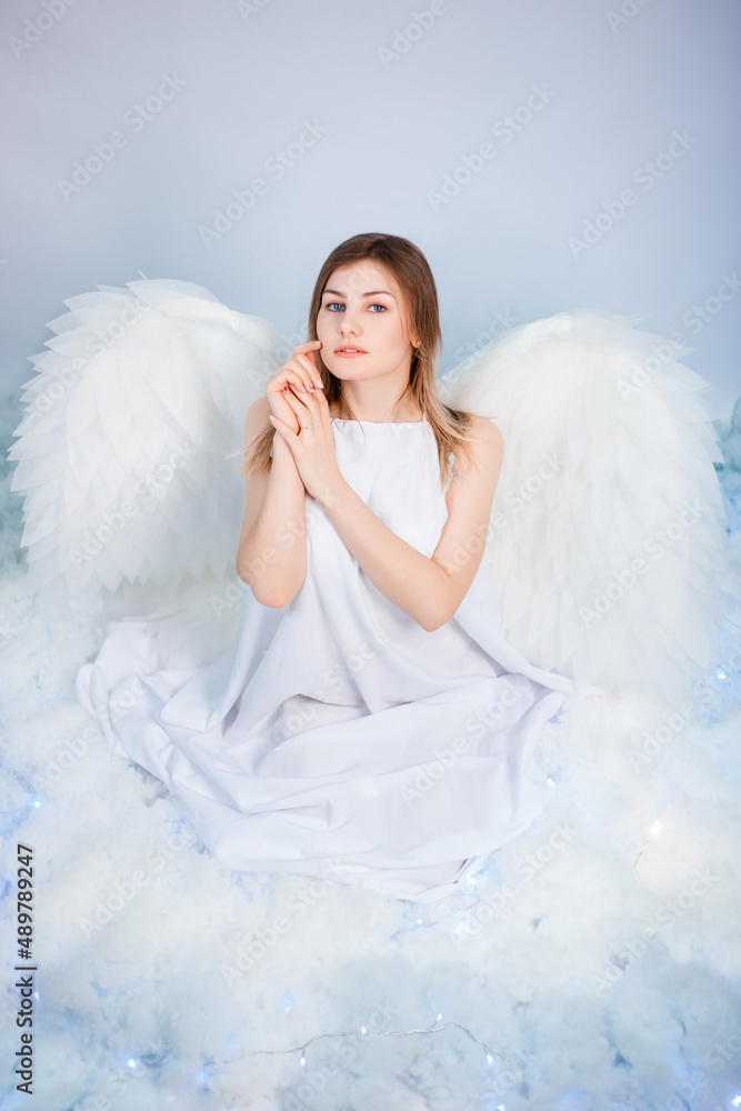 An attractive girl in a white tunic with large white wings behind her back poses while sitting in white luminous clouds. An attractive girl in a white tunic with large white wings behind her back poses while sitting in white luminous clouds.