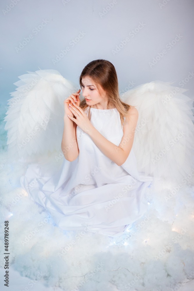 An attractive girl in a white tunic with large white wings behind her back poses while sitting in white luminous clouds. An attractive girl in a white tunic with large white wings behind her back poses while sitting in white luminous clouds.