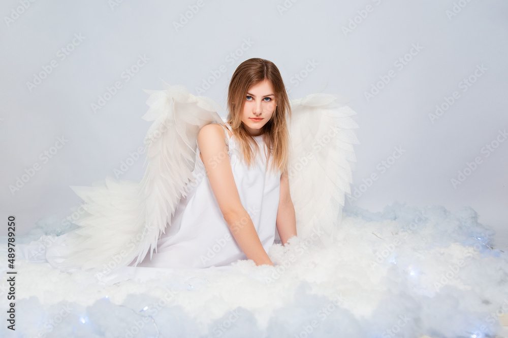 An attractive girl in a white tunic with large white wings behind her back poses while sitting in white luminous clouds. An attractive girl in a white tunic with large white wings behind her back poses while sitting in white luminous clouds.