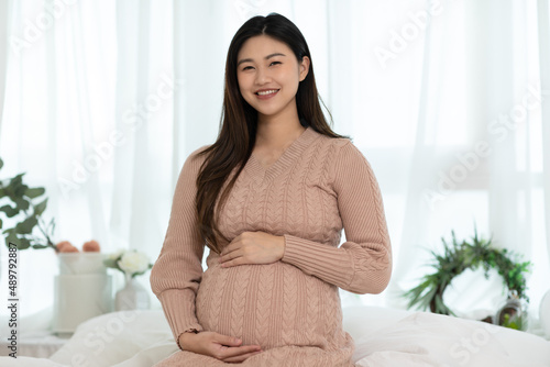 Happy Pregnant Asian Woman sitting on a bed holding and stroking her big belly at home. Pregnancy of young woman enjoying with future life relax at home. Motherhood and Pregnant Concept. Soft focus