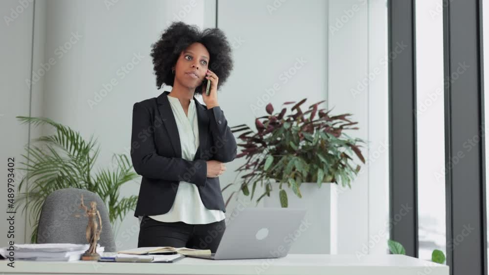 Young businesswoman or lawyer talking on phone and standing in modern office spbi. 4k video American African woman has mobile conversation and uses laptop, looks at screen and stands in light room Young businesswoman or lawyer talking on phone and standing in modern office spbi. 4k video American African woman has mobile conversation and uses laptop, looks at screen and stands in light room