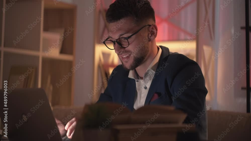 Online communication. Smiling man. Home work. Happy smart hipster guy typing laptop sitting desk with opened books in dark room interior. Online communication. Smiling man. Home work. Happy smart hipster guy typing laptop sitting desk with opened books in dark room interior.