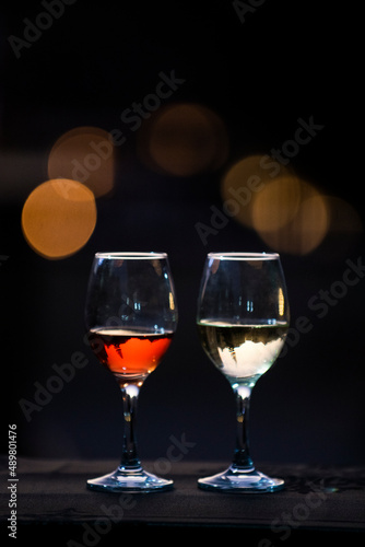 Two backlit and partially filled wine glasses with red and white wine sitting on a bar with twilight bokeh.