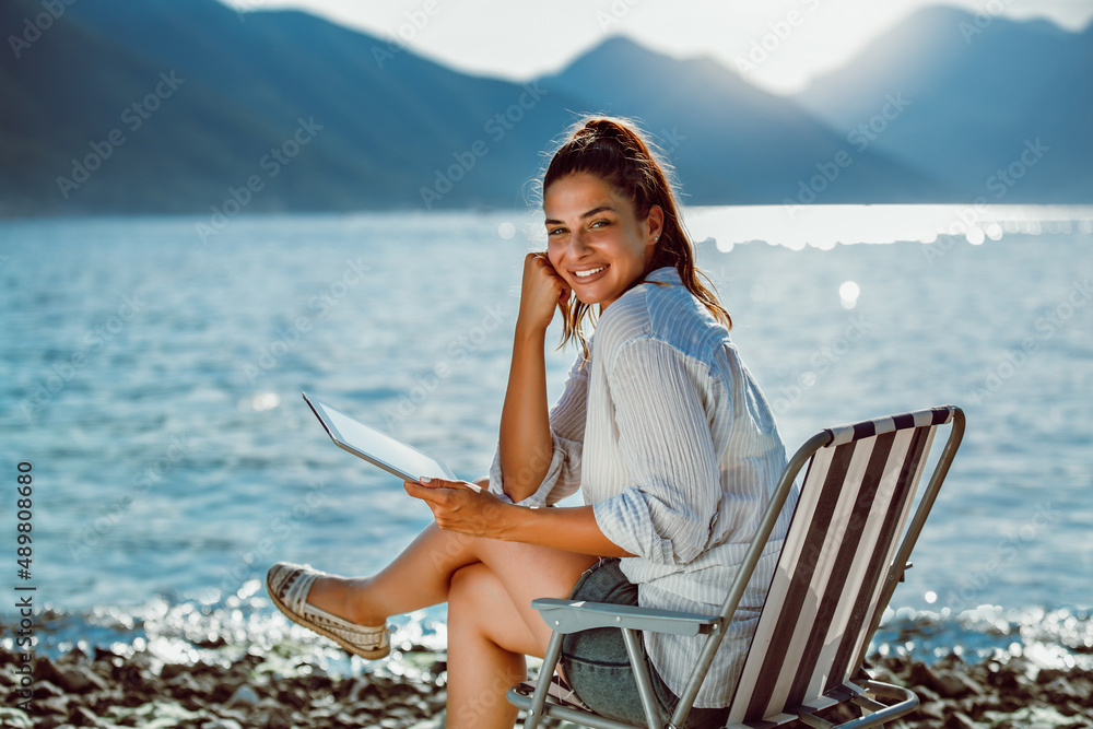 Smiling woman sitting on deck chair by the sea using tablet on a sunny day Smiling woman sitting on deck chair by the sea using tablet on a sunny day