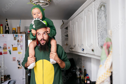 Father carrying boy on shoulders with dinosaur costume in kitchen at home photo