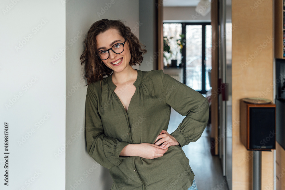 Happy young woman with hand on hip leaning on wall at home Happy young woman with hand on hip leaning on wall at home