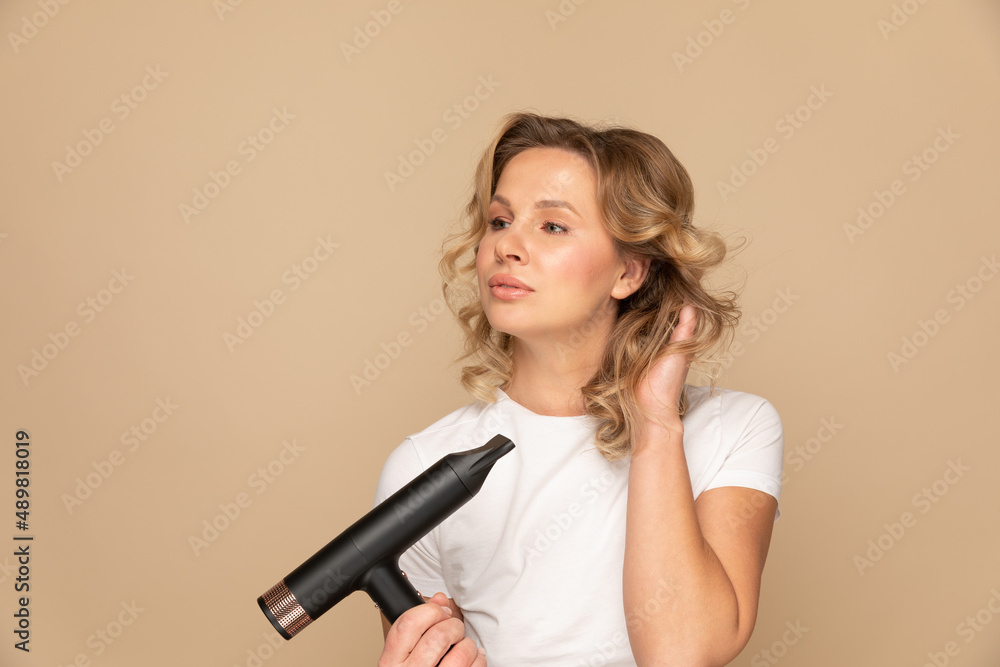 Portrait of curly blondie woman with hairdryer with cord on beige background Portrait of curly blondie woman with hairdryer with cord on beige background