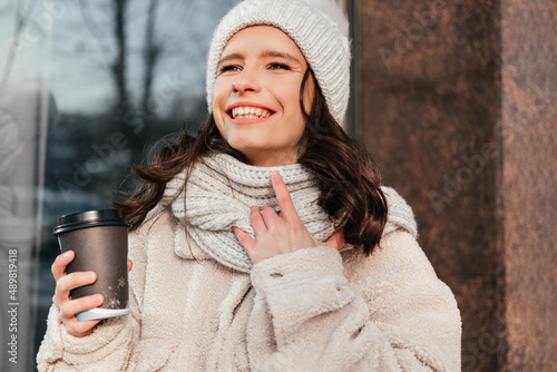 Caucasian smiling young woman in coat holding coffee cup outdoors. Woman enjoying sun