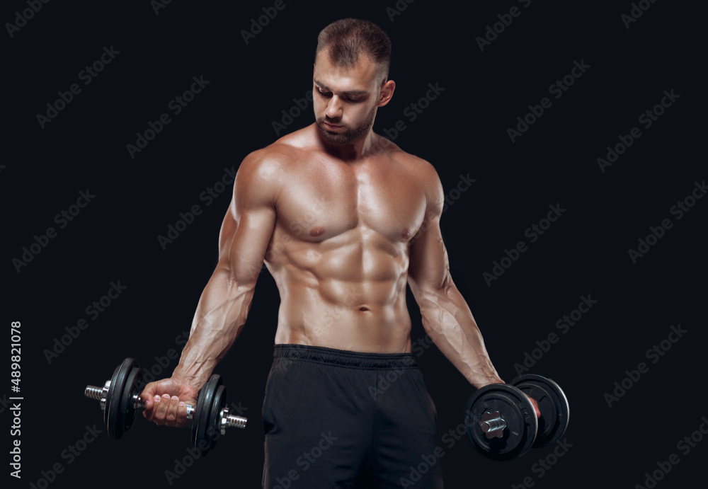 Sexy athletic man is showing muscular body with dumbbells standing with his head down, isolated over black background