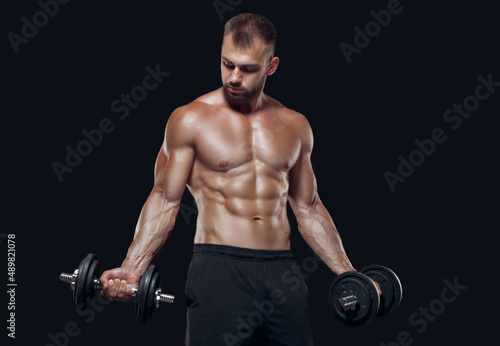 Sexy athletic man is showing muscular body with dumbbells standing with his head down  isolated over black background