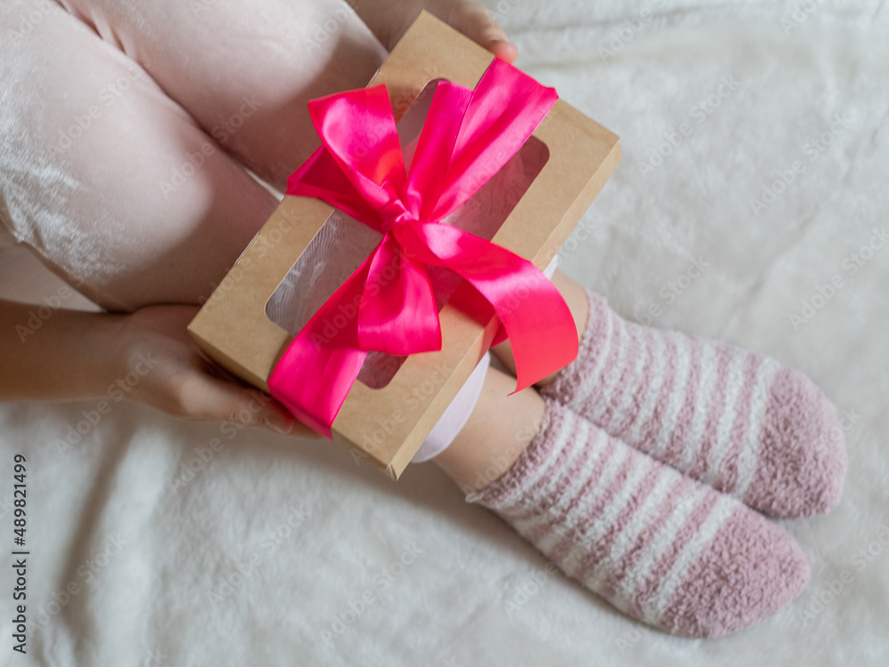 The hands of a young woman hold a box with a gift tied with a pink ribbon. A girl in light clothes sits on a white surface and holds a box with a gift. The hands of a young woman hold a box with a gift tied with a pink ribbon. A girl in light clothes sits on a white surface and holds a box with a gift.
