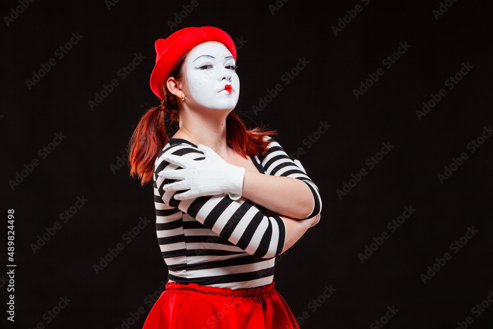Portrait of female mime artist performing, isolated on black background. Woman in striped clothes and red skirt is standing with crossed arms over her chest, a haughty look