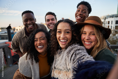 Multi-cultural group of friends taking a selfie at a rooftop party. Close up, smiles with diverse young adults.