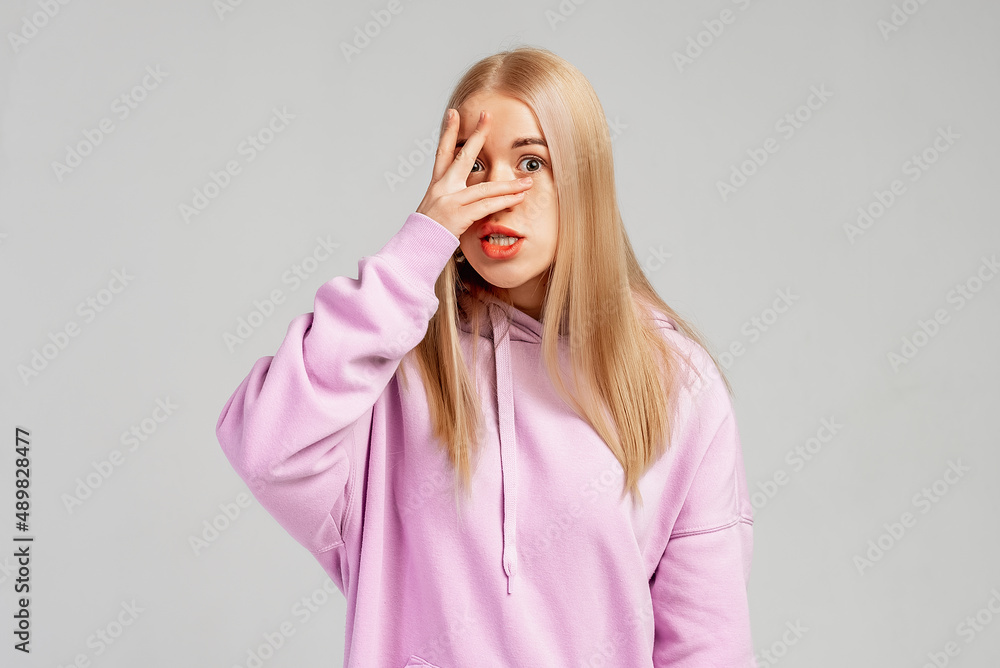 Scared blonde girl covering face, looking worried and shocked, frightened of something, wears casual clothes, stands against grey background. Body language concept Scared blonde girl covering face, looking worried and shocked, frightened of something, wears casual clothes, stands against grey background. Body language concept