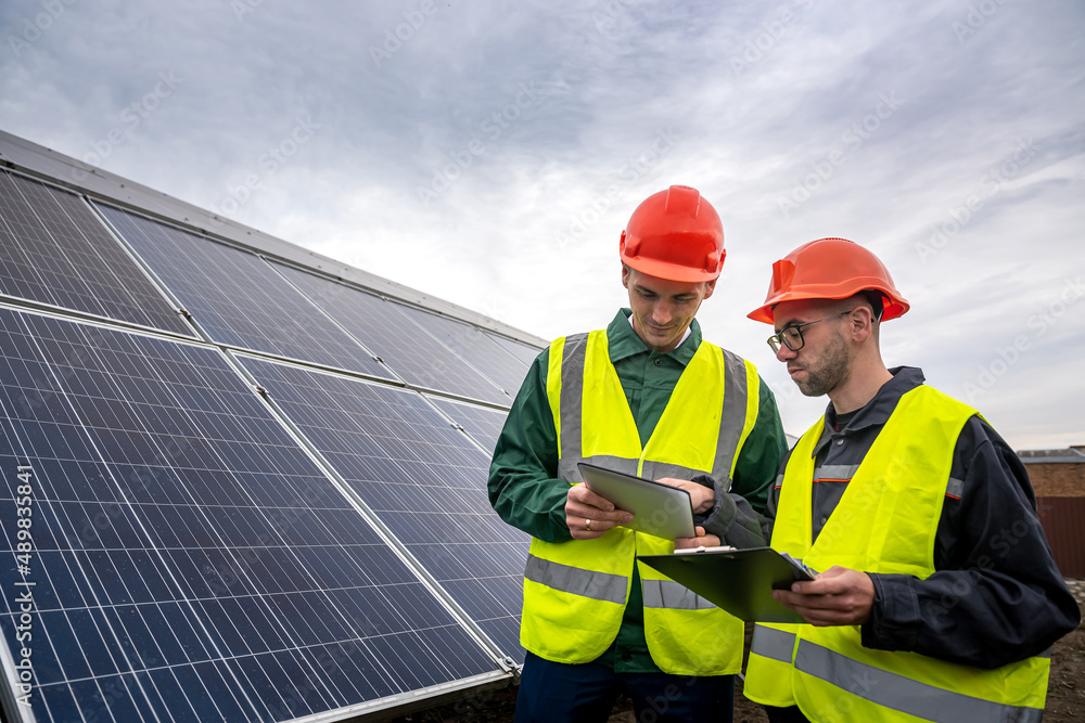 two strong skilled men in helmets and vests stand and talk in solar panels. two strong skilled men in helmets and vests stand and talk in solar panels.