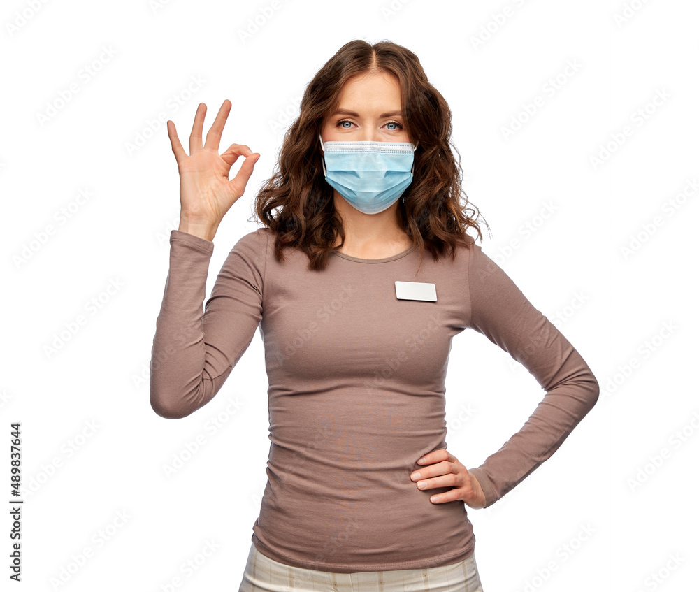 sale, shopping and pandemic concept - happy female shop assistant in medical mask with name tag showing ok gesture over white background sale, shopping and pandemic concept - happy female shop assistant in medical mask with name tag showing ok gesture over white background