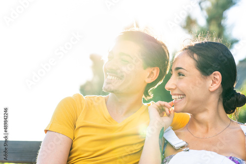 Handsome man wearing yellow tshirt and blue swimsuit sits with beautiful woman in dungarees in a park in a sunny day during holidays. Young adults couple sitting togheter surrounded by green trees