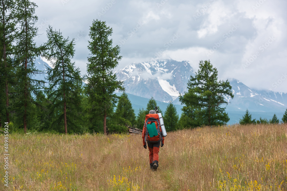 Traveling man in red with big backpack on way to snowy mountain range in low clouds. Backpacker walks through forest to large mountains under cloudy sky. Dramatic landscape with tourist in mountains. Traveling man in red with big backpack on way to snowy mountain range in low clouds. Backpacker walks through forest to large mountains under cloudy sky. Dramatic landscape with tourist in mountains.