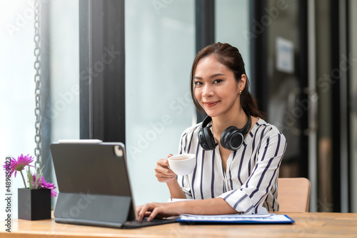 Charming young Asian businesswoman sitting smiling working drinking coffee and using laptop on the desk in the office. © amnaj