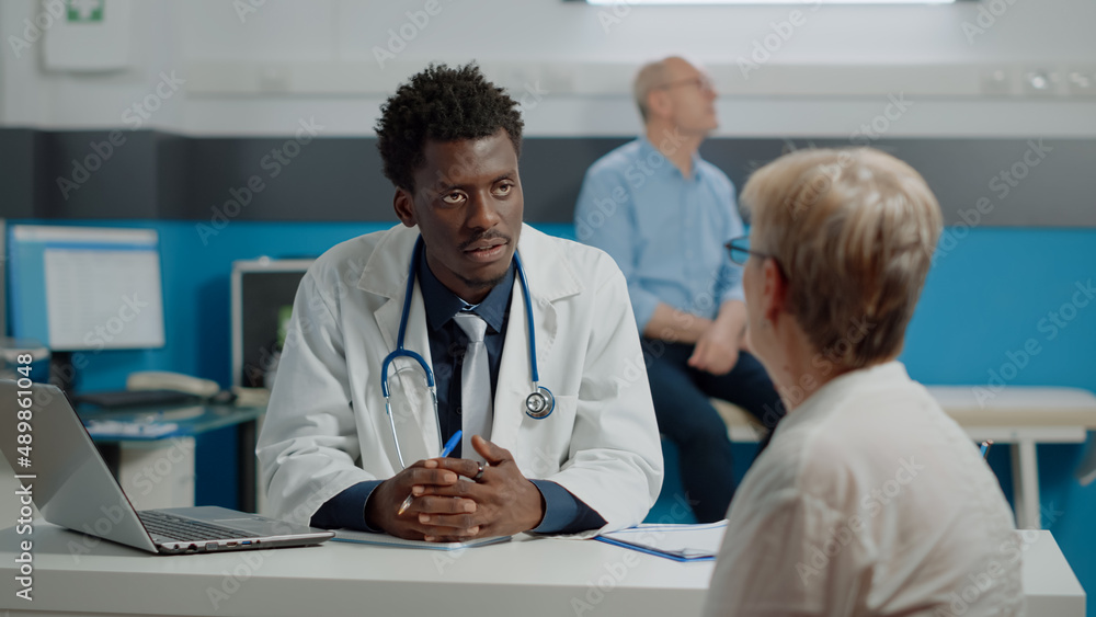 Fototapeta premium Adult specialist consulting senior woman sitting at desk writing checkup notes on files in medical office. Old patient receiving professional advice for man on bed in background