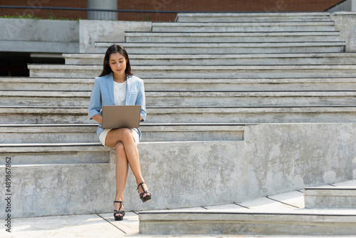 Happy business woman working on a laptop on stairs on stairway near office buildings at downtown. Confident women work online on laptop towards their goals for success at the city. © Chanakon