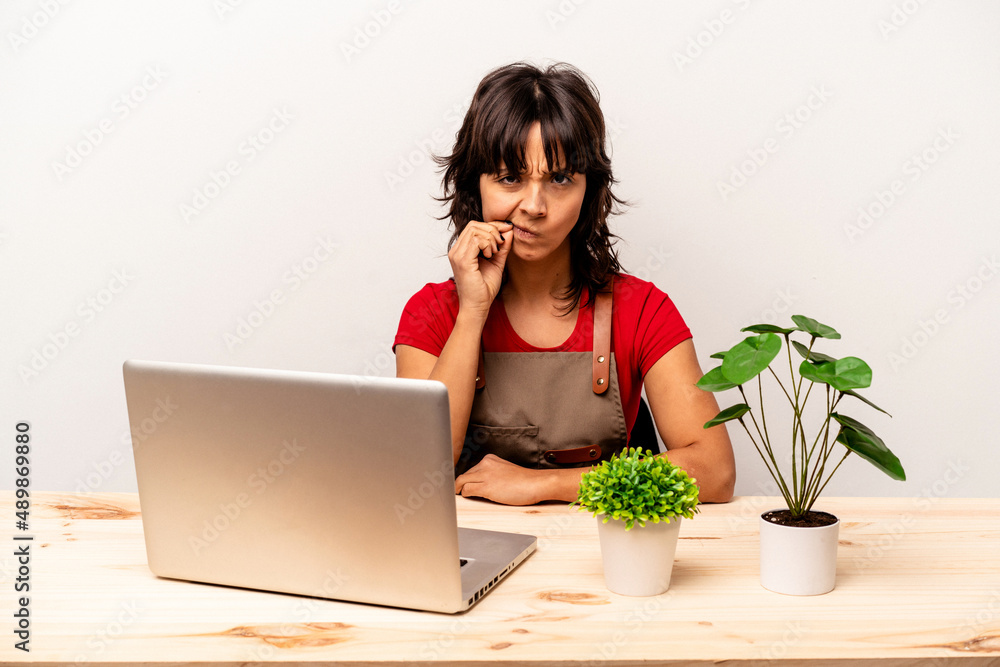 Young gardener hispanic woman sitting on a table isolated on white background with fingers on lips keeping a secret. Young gardener hispanic woman sitting on a table isolated on white background with fingers on lips keeping a secret.