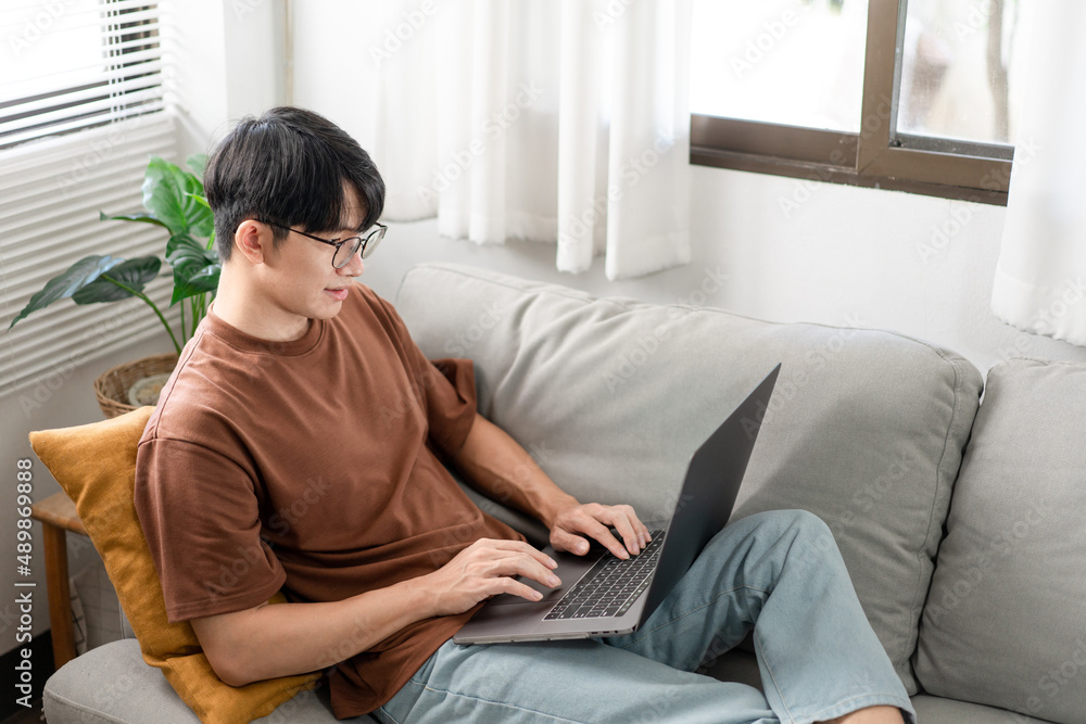 Technology Concept The man that is putting on glasses leaning his back on the dark yellow while focusing on the laptop’s screen