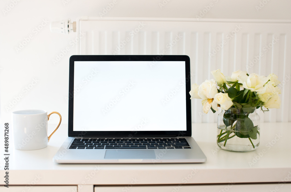 Laptop with white blank screen on white work table. White interior home office concept. Female office desk workspace with white roses and cup of coffee. Close up on screen laptop. Mock up laptop Laptop with white blank screen on white work table. White interior home office concept. Female office desk workspace with white roses and cup of coffee. Close up on screen laptop. Mock up laptop