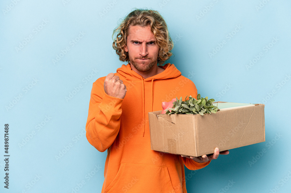 Young caucasian man making a move while picking up a box full of things isolated on blue background showing fist to camera, aggressive facial expression. Young caucasian man making a move while picking up a box full of things isolated on blue background showing fist to camera, aggressive facial expression.