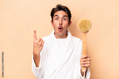 Young caucasian man wearing a bathrobe holding a back scratcher isolated on beige background having some great idea, concept of creativity.