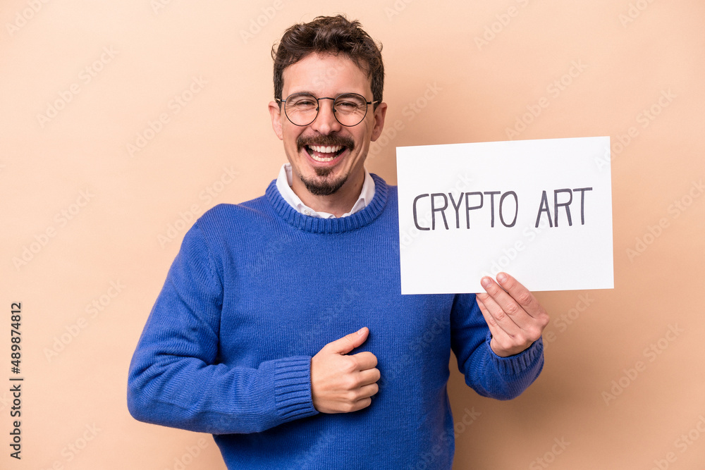Young caucasian man holding a crypto art placard isolated on beige background laughing and having fun. Young caucasian man holding a crypto art placard isolated on beige background laughing and having fun.