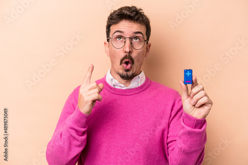 Young caucasian man holding batteries isolated on beige background pointing upside with opened mouth.