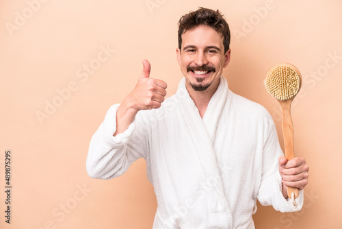 Young caucasian man holding back scratcher isolated on beige background smiling and raising thumb up