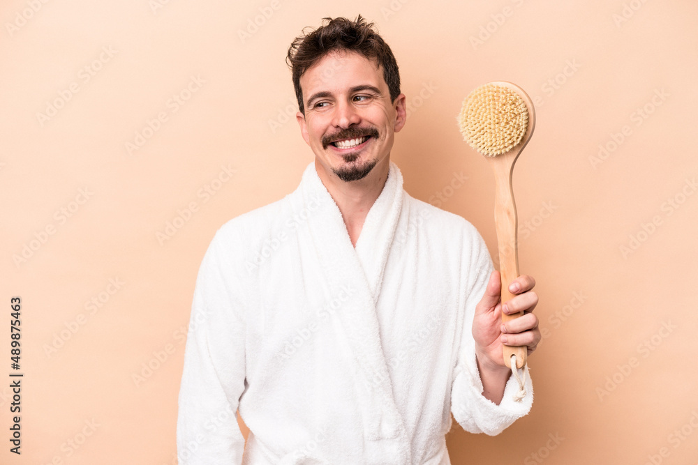 Young caucasian man holding back scratcher isolated on beige background looks aside smiling, cheerful and pleasant. Young caucasian man holding back scratcher isolated on beige background looks aside smiling, cheerful and pleasant.
