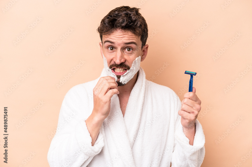 Young caucasian man wearing shaving foam and holding razor blade isolated on beige background biting fingernails, nervous and very anxious. Young caucasian man wearing shaving foam and holding razor blade isolated on beige background biting fingernails, nervous and very anxious.
