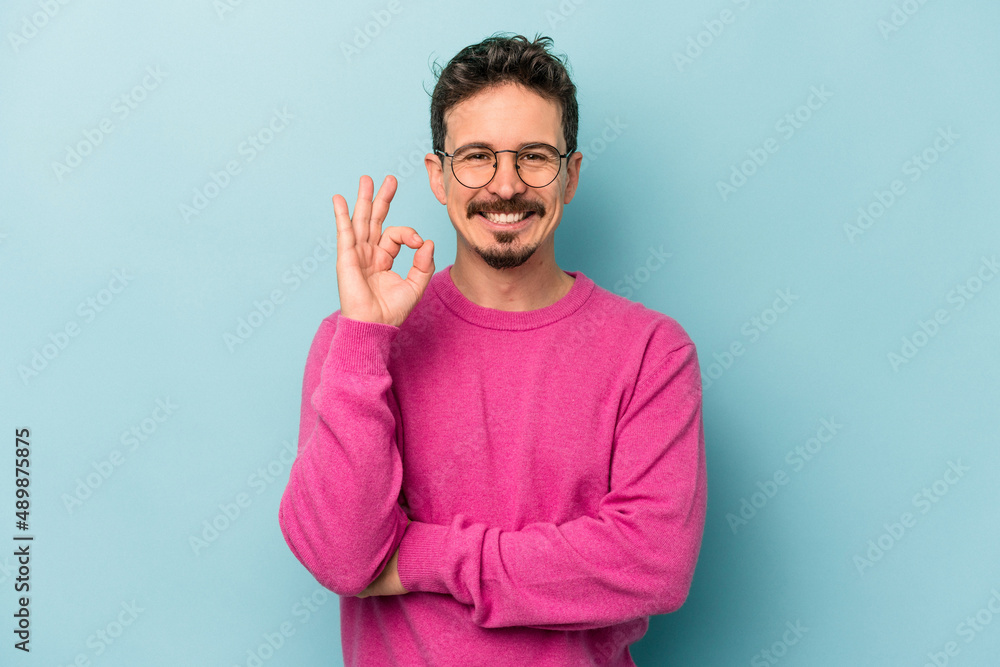 Young caucasian man isolated on blue background winks an eye and holds an okay gesture with hand. Young caucasian man isolated on blue background winks an eye and holds an okay gesture with hand.
