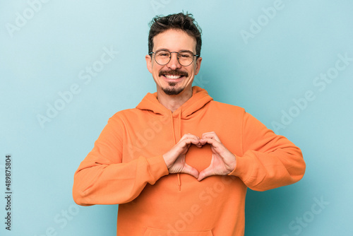 Young caucasian man isolated on blue background smiling and showing a heart shape with hands. photo