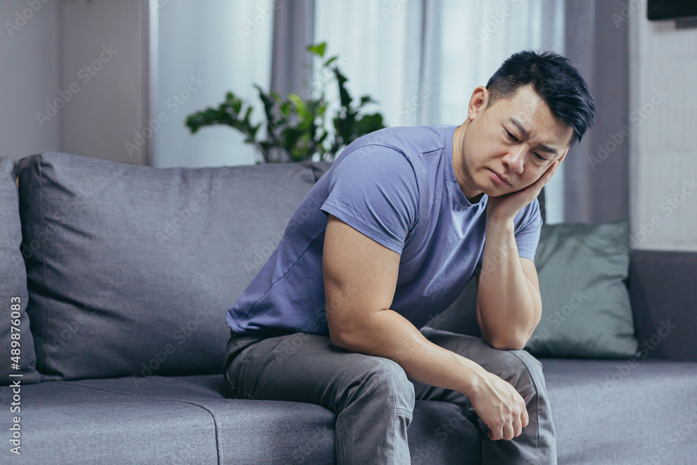 Upset Asian man at home, tired and depressed sitting on the couch in the living room