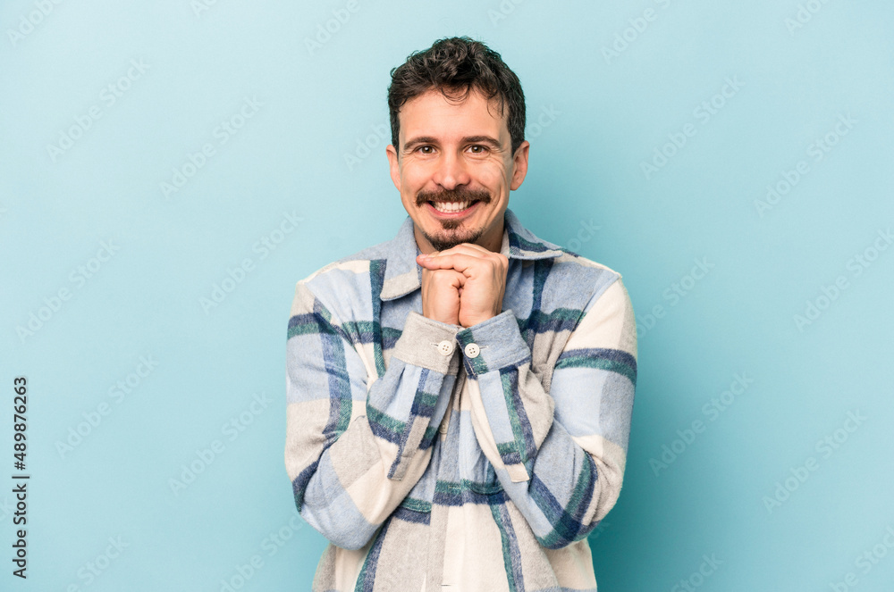 Fototapeta premium Young caucasian man isolated on blue background keeps hands under chin, is looking happily aside.