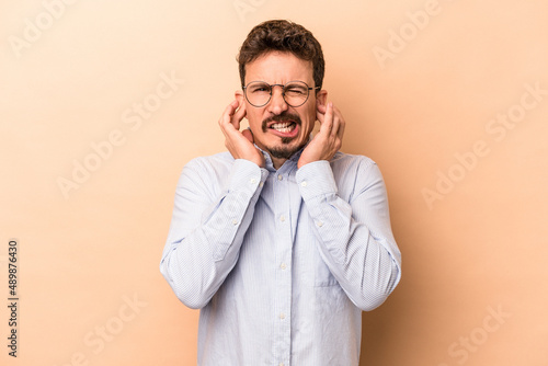 Young caucasian man isolated on beige background covering ears with hands.